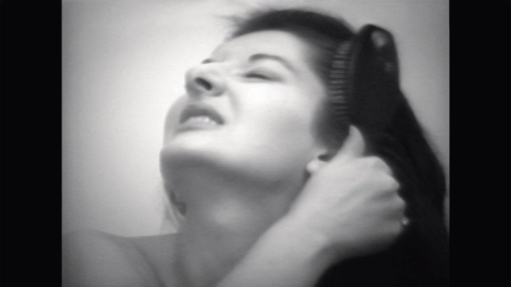 A black and white photo of Marina Abramović with her head tilted to the sky. She is baring her teeth and is dragging a brush through her long dark hair.