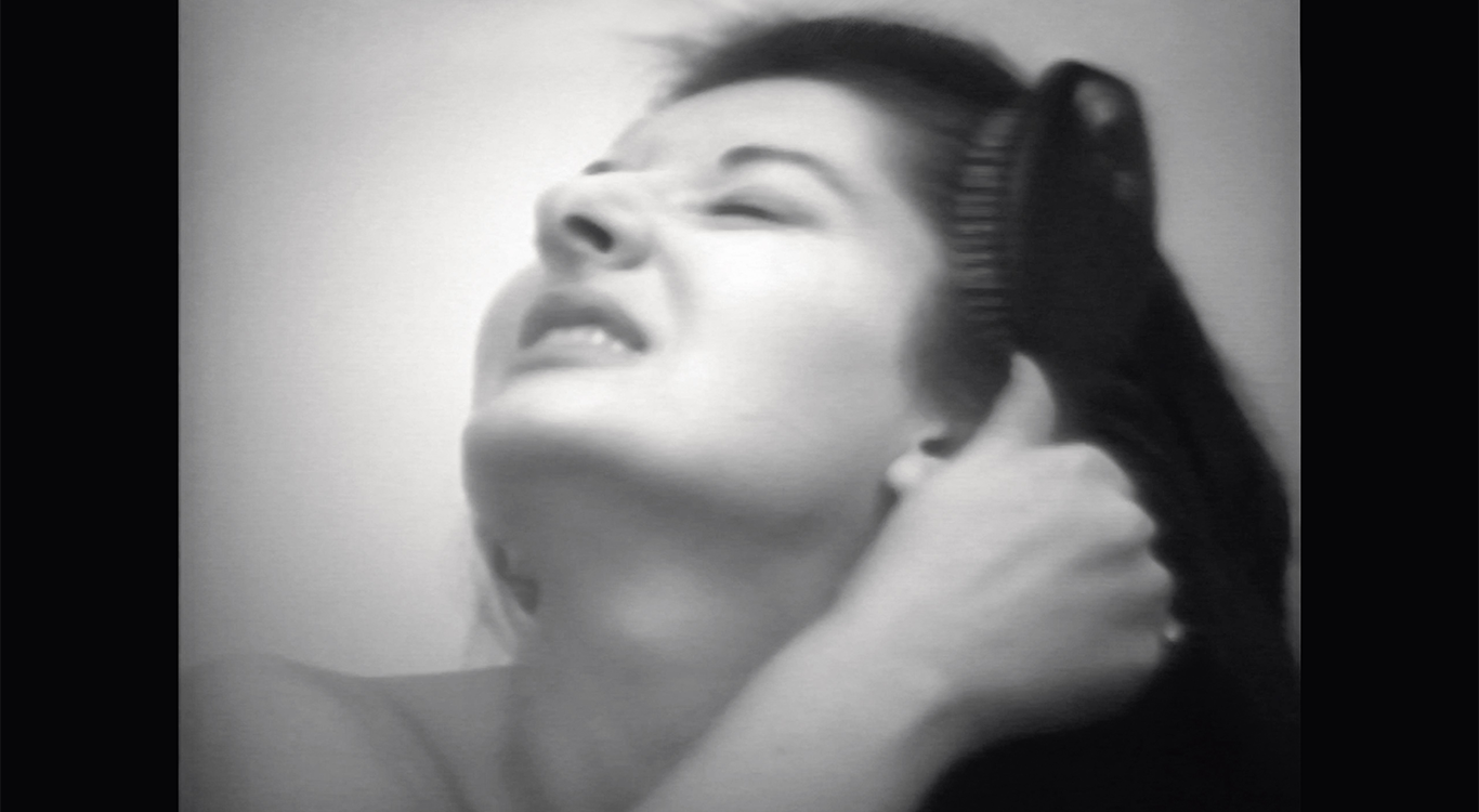 A black and white photo of Marina Abramović with her head tilted to the sky. She is baring her teeth and is dragging a brush through her long dark hair.