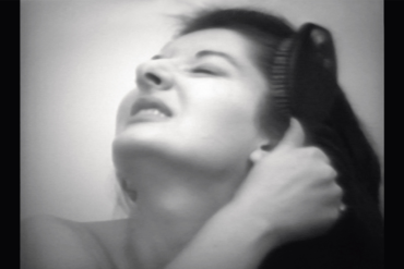 A black and white photo of Marina Abramović with her head tilted to the sky. She is baring her teeth and is dragging a brush through her long dark hair.