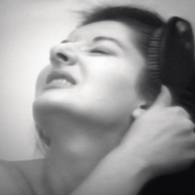 A black and white photo of Marina Abramović with her head tilted to the sky. She is baring her teeth and is dragging a brush through her long dark hair.